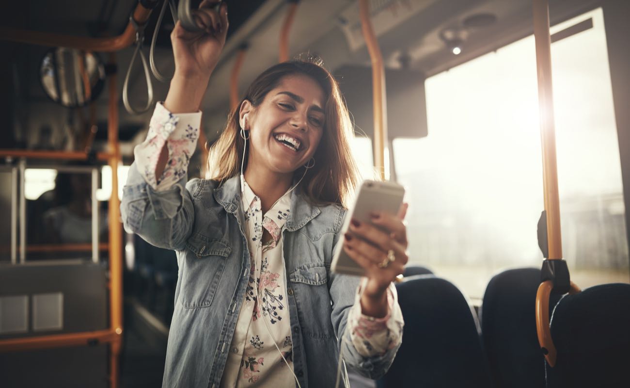  Young woman wearing earphones laughing at a text message on her cellphone while riding on a bus.