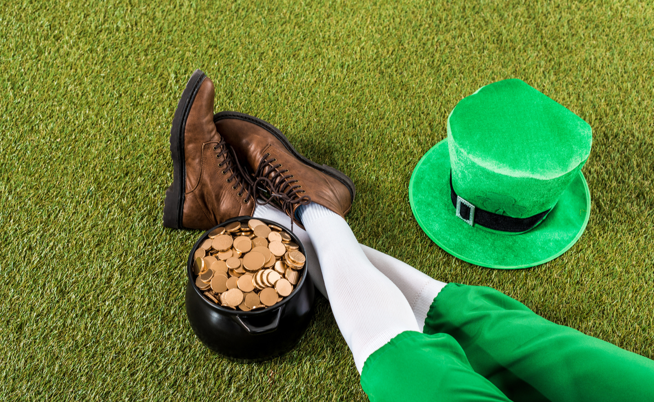 A leprechaun sitting on the grass next to a pot of gold with a top hat next to him.