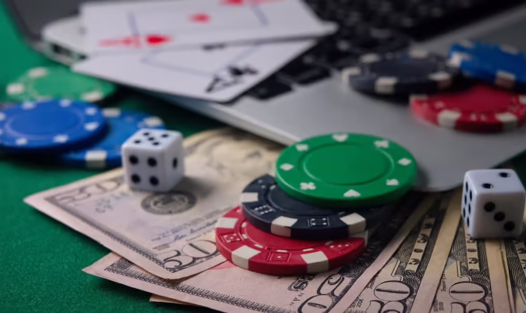 Poker chips, dice, playing cards, and U.S. currency are arranged on green felt beside a laptop.