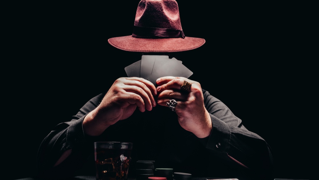 Photo of a person in black shirt and hat playing poker and holding black cards with game chips and money.