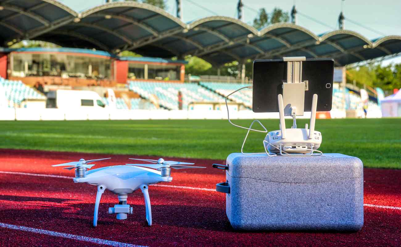 White quadcopter drone with four motors and propellers in large stadium next to carry box and remote controller with touchpad