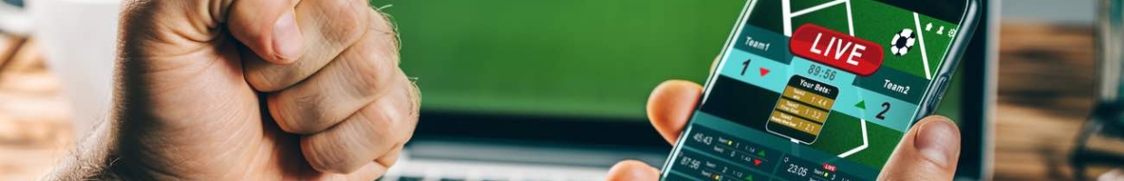 Man celebrating victory while using gambling mobile application on his phone with sports on laptop screen in the background